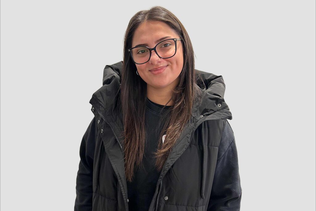 Frau mit langen braunen Haaren trägt schwarze Steppweste über schwarzem Pullover vor hellem Hintergrund.