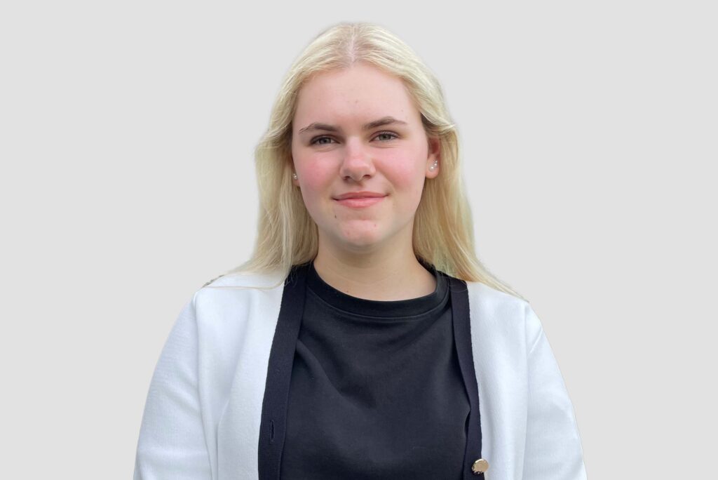 Person mit langen blonden Haaren trägt schwarzen Pullover und weiße Strickjacke vor hellem Hintergrund